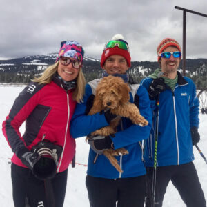 Group of nordic skiiers with a SLR camera and puppy being held in the image.