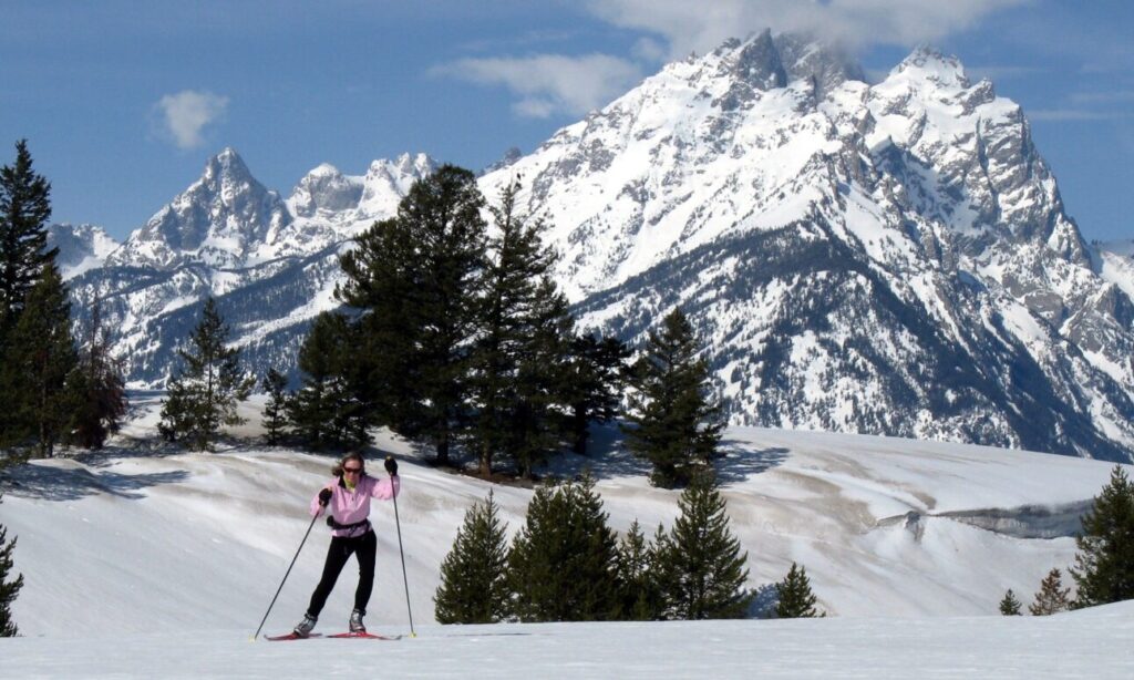 A skier in a pink top glides over the snow with the Tetons as a gorgeous backdrop
