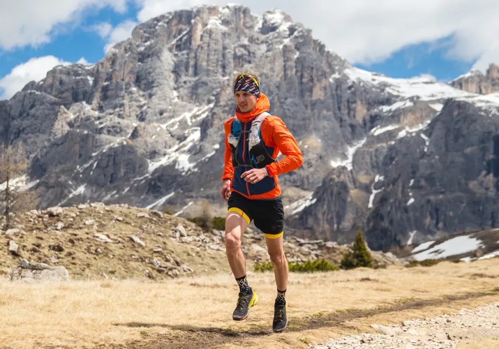 Man, Nicola Giovanelli, in orange jacket and black shorts runs in the mountains