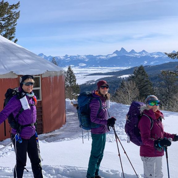 three women on cross-country skis in front of yurt with Tetons in distance