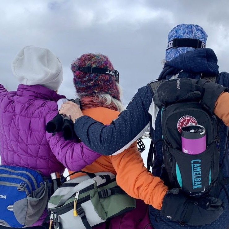 Three cross country skiers wearing winter Nordic packs with water, snacks, and emergency gear.